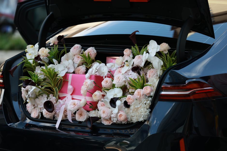 A close-up view of the open trunk of a dark-colored car filled with a vibrant floral arrangement featuring white orchids, pale pink roses, and dark purple calla lilies, all arranged around pink gift b