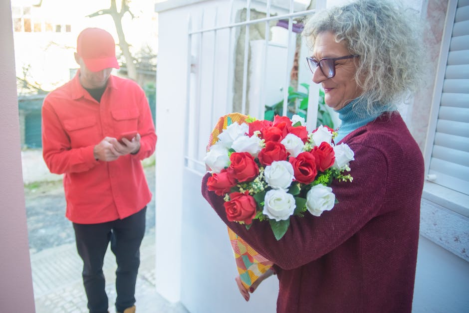 A woman with curly gray hair and glasses is holding a bouquet of fresh red and white roses wrapped in colorful paper, standing near a door in an outdoor or porch setting. She appears to be delighted a