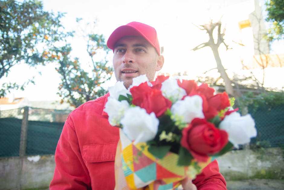 A delivery person wearing a red uniform and matching cap is holding a vibrant bouquet of fresh flowers, featuring red and white roses and white lisianthus, wrapped in a colorful paper with a yellow ri