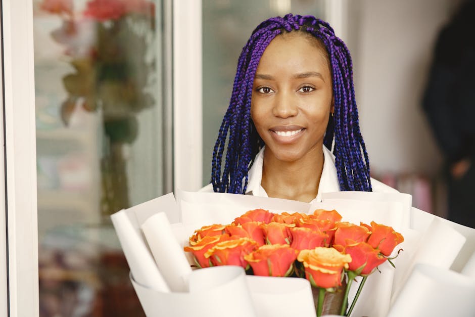 A young woman with dark skin and vibrant purple braided hair holds a large bouquet of fresh, peach and coral roses wrapped in white paper. She is smiling gently and standing indoors near a glass windo