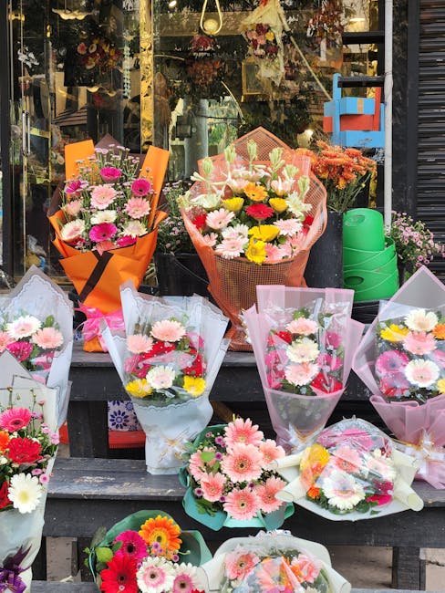 A display of various fresh flower bouquets outside a florist shop, featuring a mix of pink, white, yellow, and red daisies, chrysanthemums, and other mixed blooms. The bouquets are wrapped in colorful