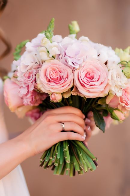 A person holding a bridal bouquet with pale pink and white roses, complemented by smaller white flowers and lush green foliage. The bouquet is arranged in a rounded, compact style with the stems wrapp