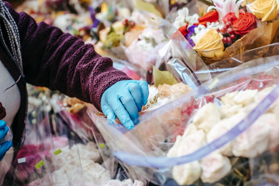 A person wearing a dark purple, textured sweater and blue rubber gloves is handling a clear plastic bag filled with various fresh flowers, including roses in shades of yellow, pink, and red, along wit