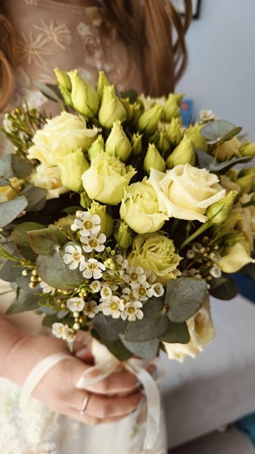 A floral bouquet featuring cream and pale yellow roses, complemented by small white daisy-like flowers and sprigs of eucalyptus with rounded, silvery-green leaves. The bouquet is held by a person wear