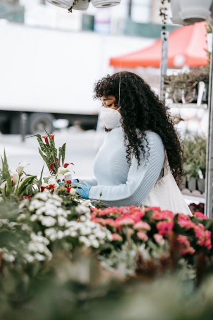 A woman with dark, curly hair wearing a white face mask, light blue sweater, and gloves is selecting fresh flowers at a flower stall. She is holding a bouquet of red and white flowers with green folia